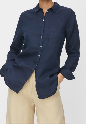 Frau trägt ein marineblaues, langärmliges Hemd mit Knöpfen und Brusttasche, kombiniert mit hoch taillierter beiger Hose, Hände in den Taschen.