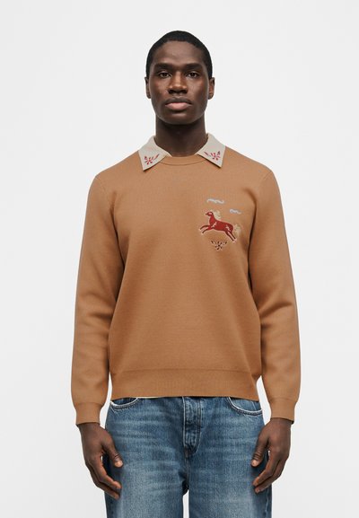 Άνδρας που φοράει πουλόβερ σε ταμπά χρώμα με κεντημένο κόκκινο άλογο και εκρού γιακά με κόκκινα λουλουδάτα σχέδια, σε συνδυασμό με μπλε τζιν.