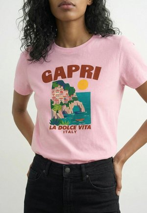 Vrouw die een roze T-shirt draagt met een kleurrijk kusttafereel van Capri, een gele zon en de tekst "CAPRI LA DOLCE VITA ITALY" voorop.