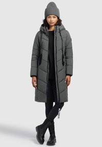 Manteau long matelassé gris foncé avec des coutures en zigzag, capuche et poches noires, assorti à un bonnet et des bottines noires.