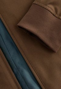 Braune Stoffjacke mit glattem Futter. Verfügt über einen Reißverschluss und weiche, gerippte Bündchen. Strukturierte Außenseite mit einem dezenten Muster im Futter.
