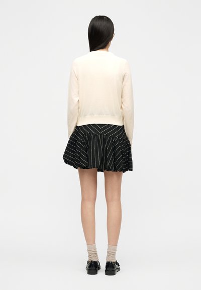 Γυναίκα με μακριά σκούρα μαλλιά φοράει μπεζ πουλόβερ, μαύρη μίνι φούστα με άνοιγμα και λευκές ρίγες, μπεζ κάλτσες και μαύρα παπούτσια τύπου λόφερ, γυρισμένη προς τα πίσω.