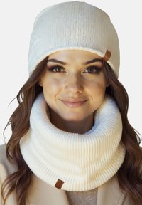 Hvid strikket hue med opbuk og matchende tyk ribstrikket halsedragt. Begge har et lædermærke, der giver et hyggeligt, tekstureret udseende.