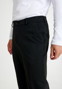 Zwarte, gestructureerde broek met een slim fit, voorzien van een knoopsluiting en riemlussen, gedragen met een wit overhemd met lange mouwen.