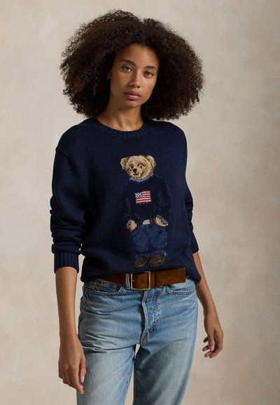 Young woman with curly hair wearing a navy blue sweater featuring a teddy bear with an American flag, paired with light blue jeans and a brown belt.
