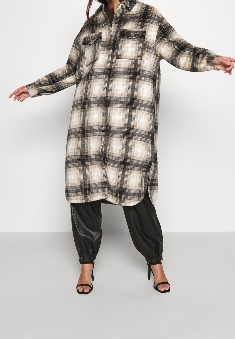 Femme portant une longue robe-chemise à carreaux beige et noire boutonnée, sur un pantalon jogger en cuir noir et des sandales à talons hauts noires à bout ouvert.