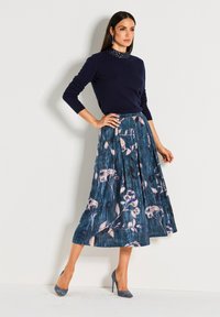 Frau, die in einem marineblauen Pullover, einem blumigen, plissierten blauen Mididress und passenden blauen High Heels vor einem schlichten weißen Hintergrund posiert.