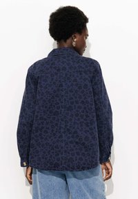 Veste en denim bleu marine avec un motif léopard foncé, poignets à boutons et une coupe légèrement décontractée. Texture lisse et design de col classique.