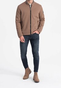 Brun zip-up jakke med en tekstureret overflade, stående krave og sidelommer med lynlås, parret med mørke jeans og brune ankelstøvler.