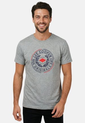 Homme souriant avec les cheveux courts et foncés, portant un t-shirt gris Lee Cooper Originals avec un logo circulaire, debout devant un fond uni clair.
