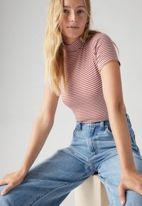 Randig t-shirt i röd och vit, hög hals, korta ärmar; tillsammans med ljusblå högmidjade jeans. Sitter på en pall.
