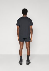 Schwarzes T-Shirt und Shorts, schlichtes Design, entspannte Passform. Schwarze Sneakers, knöchellange Socken mit Logo, stehend vor einem schlichten Hintergrund.