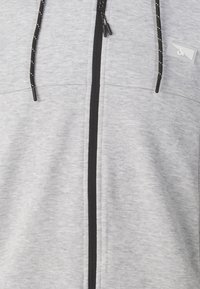 Sudadera con cremallera gris que presenta una textura suave, cremallera negra y cordones, con una pequeña etiqueta de logo en el pecho.