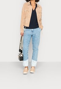 Beige mocka jacka med två framfickor, marinblå stickad tröja, ljusblå jeans med uppvikta fållar, beige slip-on skor och en liten svart axelväska.