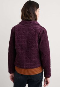 Fioletowa kurtka z korduroju o diamentowym pikowanym wzorze, szerokim kołnierzu i pomarańczowym ściągaczu. Model pokazany z tyłu, z rozpuszczonymi włosami.