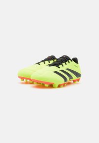 Botas de fútbol verde lima con acentos negros, parte superior texturizada y suelas naranjas brillantes. Presenta tres franjas negras y sistema de cierre con cordones.