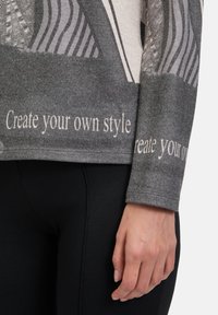 Grauer Strickpullover mit abstrakten Mustern, dem Text "Kreiere deinen eigenen Stil" in Hellgrau und einer lockeren Passform. Schwarze Leggings sind darunter sichtbar.