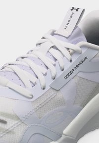 Κοντινή εικόνα λευκού αθλητικού παπουτσιού Under Armour με υφασμάτινη υφή, πυκνά λευκά κορδόνια και γλωσσίδα με επωνυμία "Reign X".