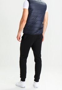 Laivaston sininen puffer-liivi, jossa vaakasuorat tikkauskuviot, yhdistetty mustiin collegehousuihin. Valkoiset tennarit viimeistelevät asun. Neutraali tausta.