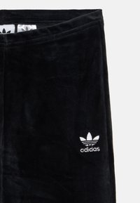 Svarte velour joggebukser med elastisk midje og hvit brodert Adidas trefoil-logo på venstre ben. Myk tekstur og minimalistisk design.