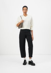 Pull à col en V crème avec ourlet côtelé, jean noir taille haute à jambes larges et chaussures à plateforme noires. Un petit sac à main est tenu dans une main.