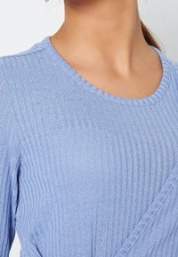 Haut côtelé bleu clair avec un col rond. Le tissu a un éclat subtil. Présente un design croisé sur le devant, mettant en valeur la texture.