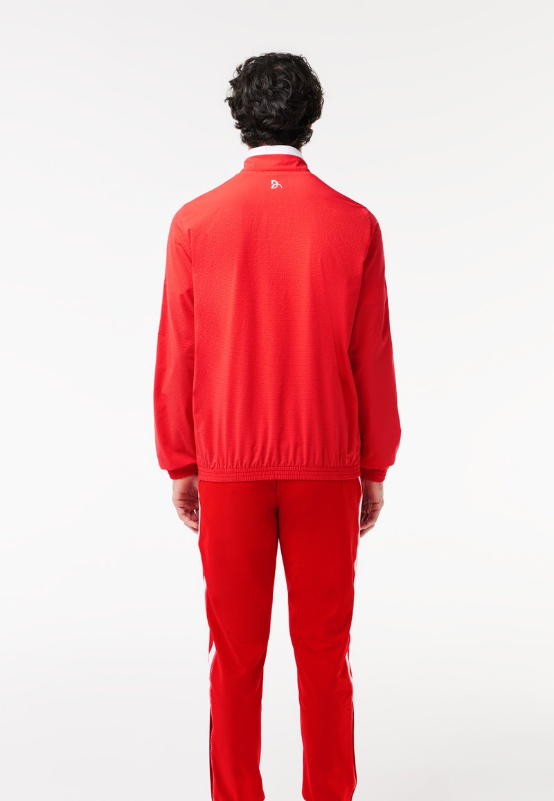 Veste de sport rouge avec une finition texturée, poignets élastiques et taille élastique. Détail de logo notable près du col. Associée à un pantalon rouge.
