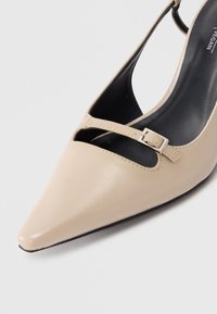 Beige vegan leather high-heeled shoe featuring a pointed toe, single strap with a rectangular silver buckle, and an open back design.