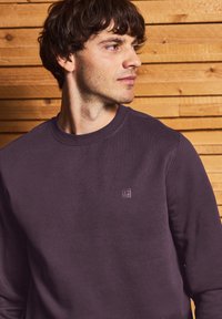 Sudadera morada con cuello redondo y un pequeño logo en el pecho. Tejido suave, mangas largas y un corte relajado. Fondo de madera.