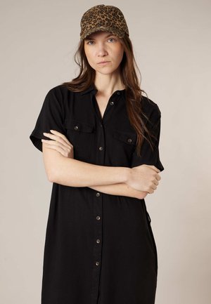 Femme les bras croisés, portant une robe noire à boutons et une casquette de baseball à imprimé léopard, sur un fond clair uni.