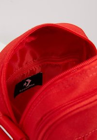 Červená taška na zip otevřená, aby odhalila vnitřní kapsu s černou nášivkou značky Converse uvnitř.