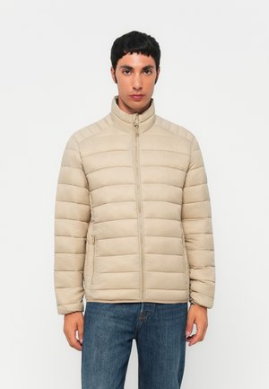 Homme portant une veste beige matelassée zippée avec poches latérales et un jean bleu, debout devant un fond blanc uni.