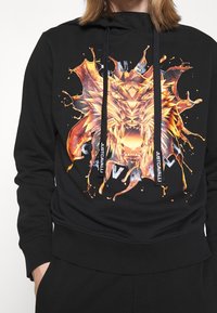 Schwarzer Hoodie mit einem lebendigen, orange-gelben Löwengrafik, schwarzen Zugbändern und dem aufgedruckten Markennamen an den Enden. Weich im Stoff.