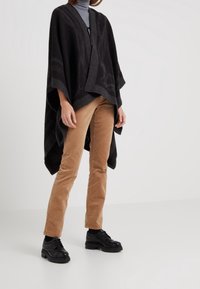 Schwarzer Poncho mit strukturiertem Muster, offener Vorderseite und weiten Ärmeln. Kombiniert mit hellbraunen Cordhosen und schwarzen Schnürschuhen.