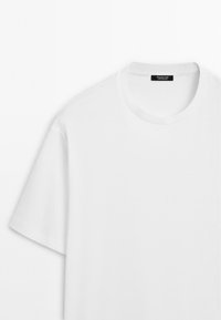 Lys hvid T-shirt med korte ærmer og rund hals, lagt fladt ud med et synligt sort mærke ved halsudskæringen.