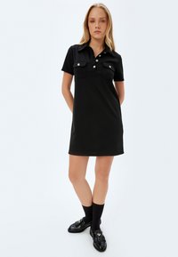 Abito stile polo nero realizzato in tessuto liscio. Presenta due tasche sul petto, cinque bottoni sul colletto e maniche corte. Indossato con scarpe e calzini neri.