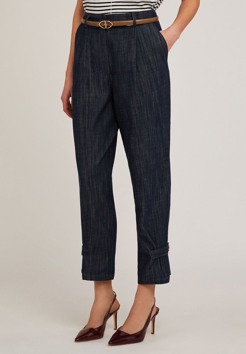 Pantalones de tiro alto, a rayas en azul oscuro, con pliegues, bolsillos laterales y de longitud hasta el tobillo, combinados con un cinturón marrón y zapatos de tacón burdeos.