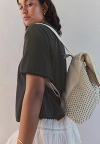 Lekki plecak w kolorze kremowym z teksturowanym splotem. Modelka nosi ciemny t-shirt z krótkim rękawem i falbaniastymi brzegami, w zestawieniu z marszczoną białą spódnicą.
