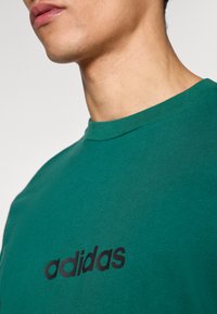 Zelené bavlnené tričko s okrúhlym výstrihom a čiernym logom adidas potlačeným na prednej strane. Hladká textúra a priliehavý strih.