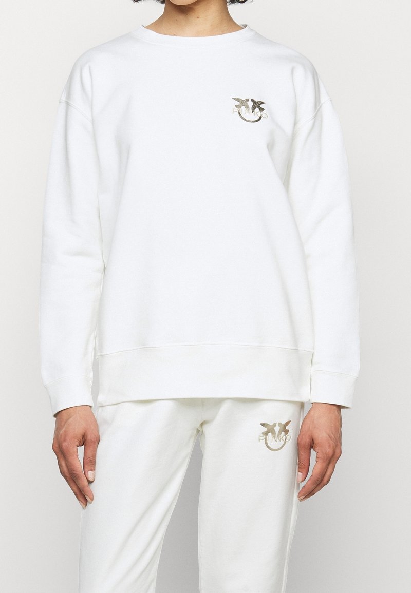 Valkoinen puuvillaneule, jossa on roikkuvat olkapäät ja leveä pyöreä kaula-aukko. Vasemmassa rinnassa on kultainen logo. Yksinkertainen, rento muotoilu.