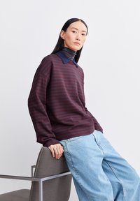 Langärmliges Sweatshirt in Kastanienbraun und Marineblau mit Kragen, kombiniert mit hellblauen weit geschnittenen Jeans, die auf einem Stuhl ruhen.