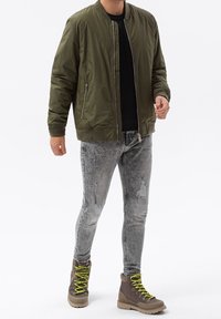 Olivgrön bomberjacka, svart skjorta, grå slitna skinny jeans och bruna vandringskängor med ljusgröna snören.