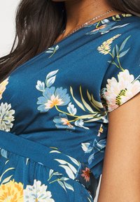 Nainen, joka on pukeutunut teal-väriseen kukkamekkoon, jossa on lyhyet hihat, ja hänellä on päällä hopeaketju. Hänet kuvataan ylävartalosta, ja hänellä on pitkät tummat hiukset.