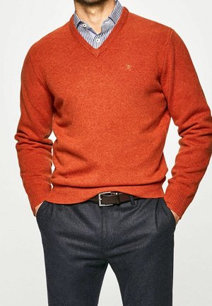 Homme portant un pull col V orange rouille sur une chemise bleue rayée avec un pantalon gris foncé et une ceinture noire, mains dans les poches, sur fond uni.