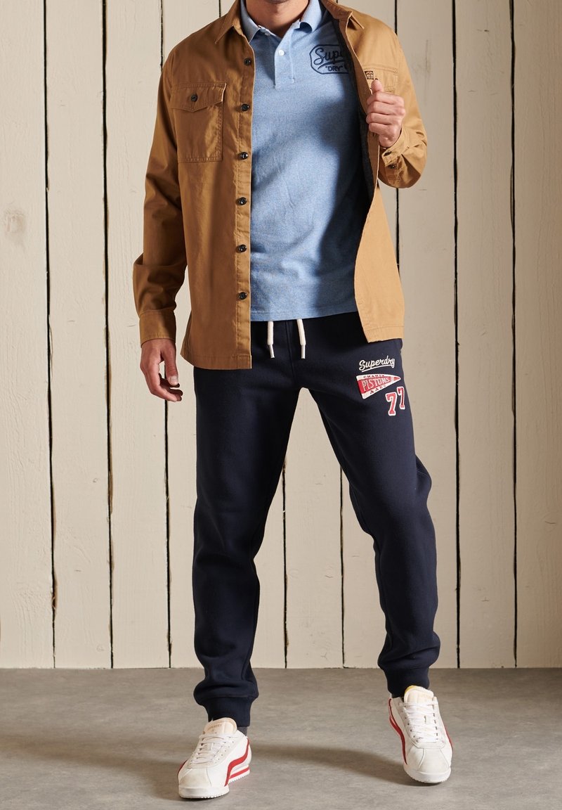 Chemise à boutons marron sur un polo bleu clair, pantalon de jogging navy avec des accents rouges et blancs, et baskets blanches avec des rayures rouges.