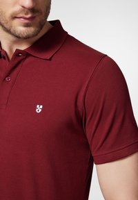 Bordowa koszulka polo wykonana z teksturowanej tkaniny, z kołnierzykiem, dwoma guzikami i małym białym logo na lewej piersi.