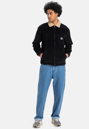 Black corduroy jacket with a beige faux fur collar, zip closure, chest pocket, worn with light blue loose-fitting jeans and gray sneakers.