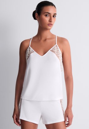 Jeune femme aux cheveux foncés attachés en chignon, portant un haut blanc sans manches et un short avec des panneaux latéraux en dentelle transparente, debout devant un fond uni.