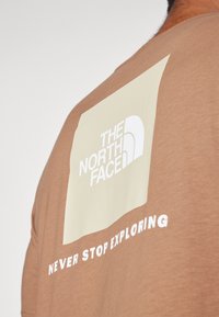 Camiseta de manga corta marrón con un gran gráfico crema que presenta el logo de "THE NORTH FACE" y el texto "NEVER STOP EXPLORING" en blanco.