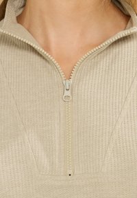 Primer piano di un top in tessuto beige a costine con una grande cerniera in plastica parzialmente chiusa e un colletto visibile attorno al collo.